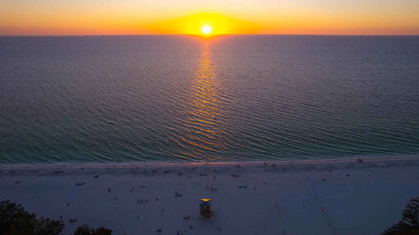 Aerial view of a sunset over the Gulf of Mexico with a wide white sand beach, a lifeguard tower, and people below.