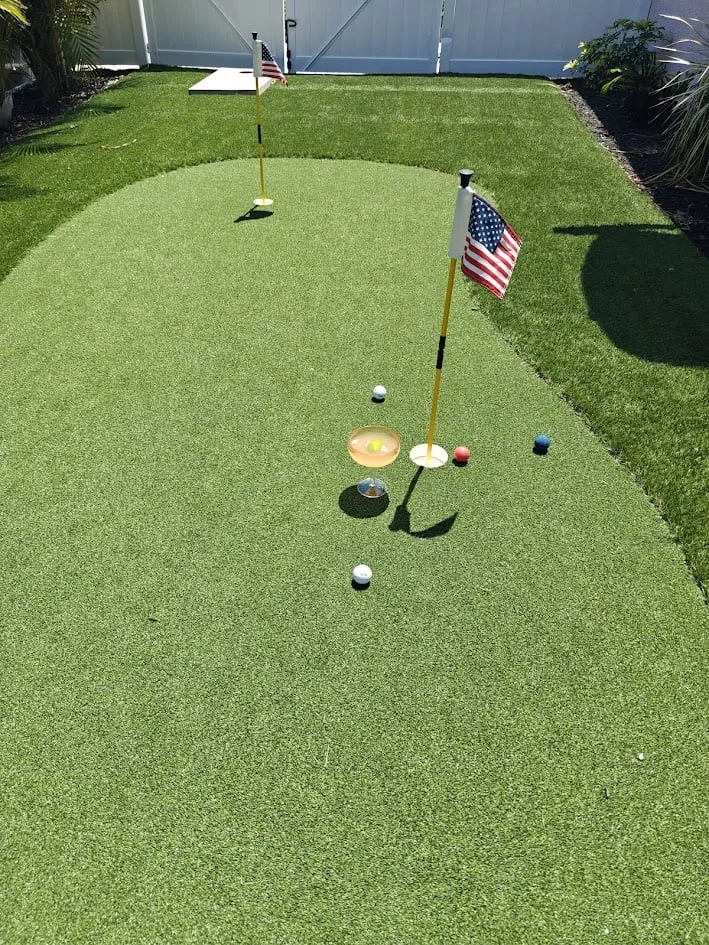 A private backyard putting green with two holes featuring American flags, golf balls, and a margarita in a coupe glass.