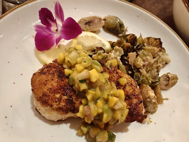 A seared grouper fillet topped with mango salsa and a purple orchid garnish, served with roasted brussels sprouts.